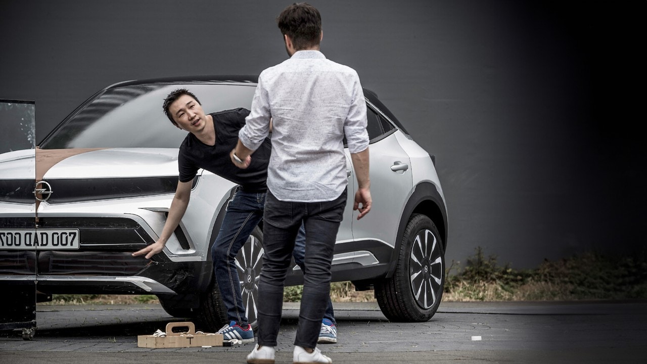 Ein junger Mann erklärt einer anderen Person etwas und deutet auf die Front eines Opel-Fahrzeugs